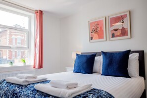 A neatly made bed with white and blue bedding, a window with a view of buildings, and two framed artworks on the wall. - Host Stay Bromyard Flat 1 (Worcester)