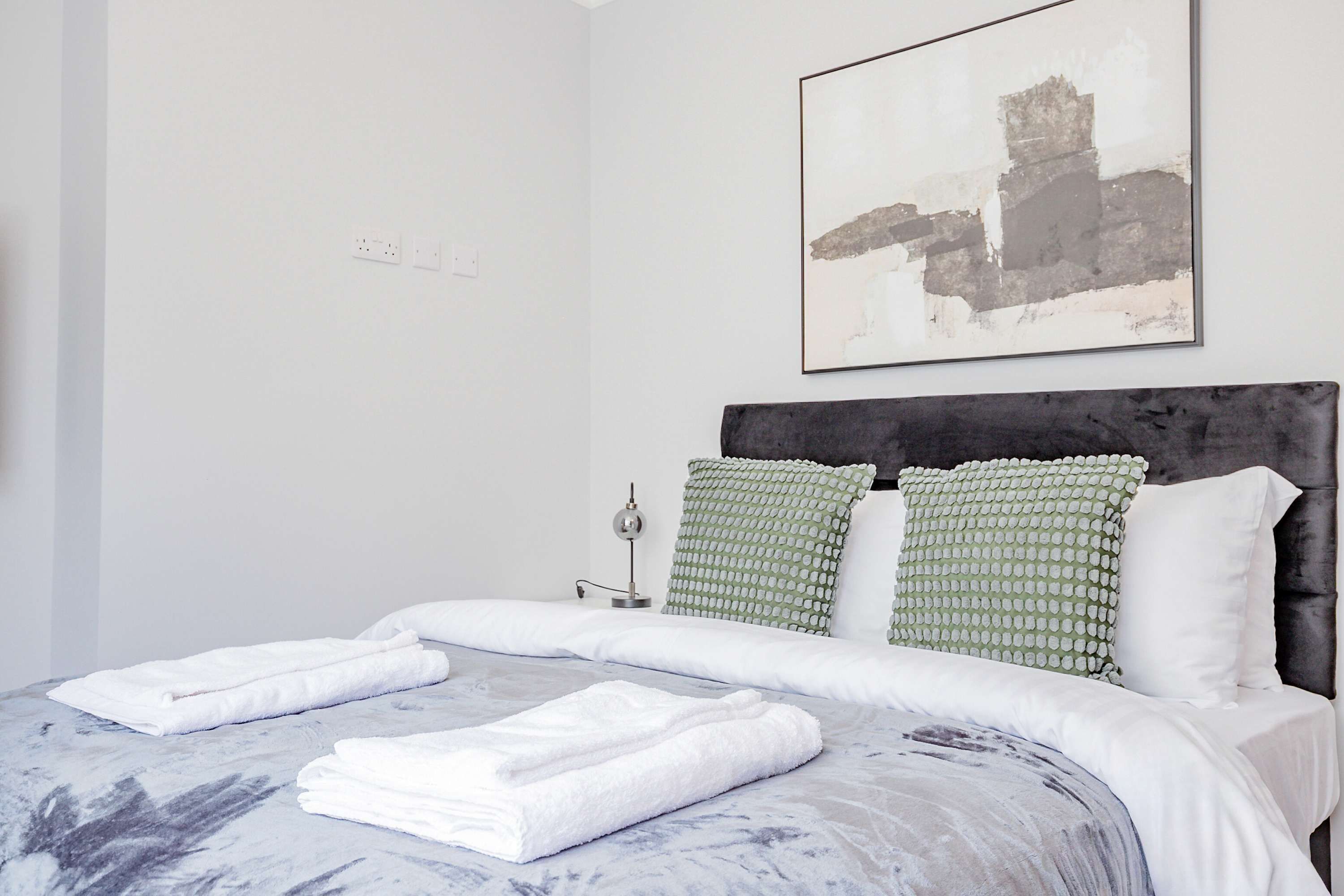 A neatly made bed with white linens and towels, a modern headboard, and a minimalist abstract painting on the wall.