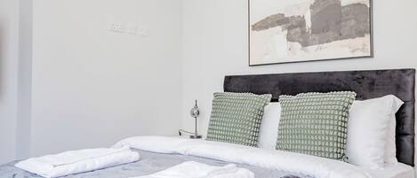 A neatly made bed with white linens and towels, a modern headboard, and a minimalist abstract painting on the wall.