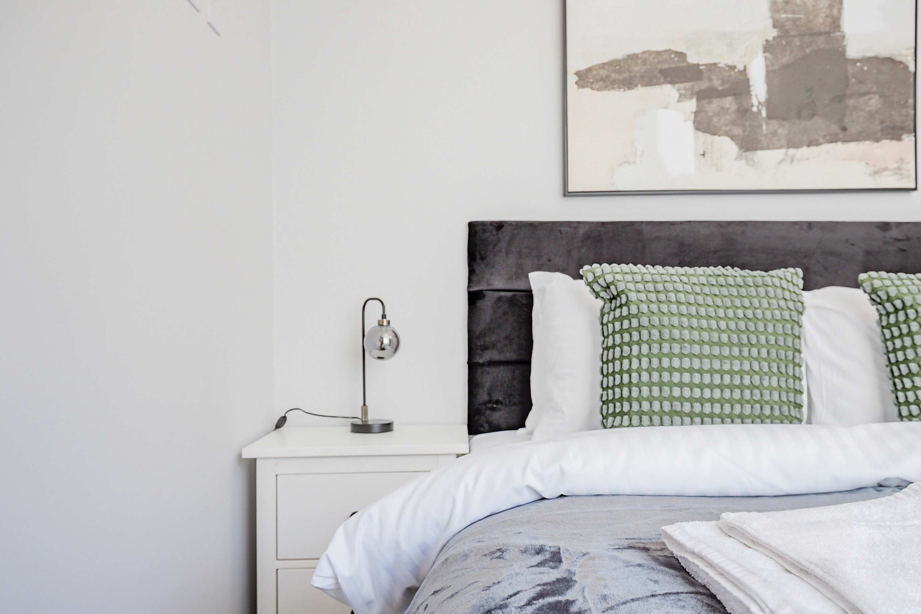 A neatly arranged bedroom with a bed, pillows, a bedside table with a lamp, and a wall-mounted artwork.