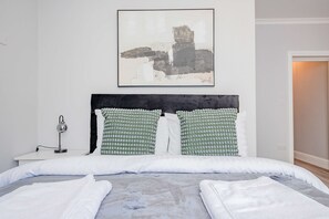 A neatly made bed with white bedding and green patterned pillows, a bedside table with a lamp, and a large abstract painting on the wall. - Host Stay Bridge Street Apartment (Evesham)