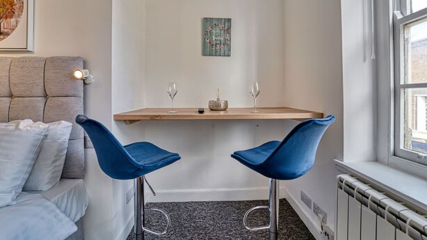 A corner room with a bed, a small table with two chairs, a decorative wall piece, and a window.