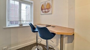 Ett litet rum med ett fönster, ett träbord, två blå barstolar och en inramad tavla på väggen.
