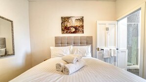 Ett hotellrum med en stor säng, en tavla som föreställer en stadsbild, ett badrum med dusch och en dörr som leder ut till en balkong.