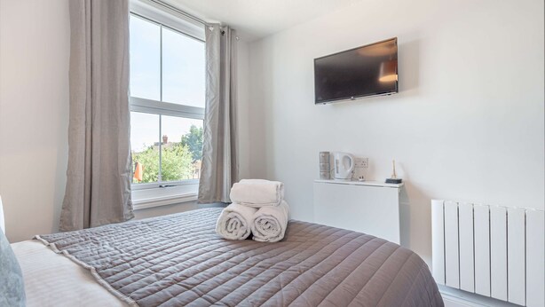 A hotel room with a large bed, a flat-screen TV mounted on the wall, a window with curtains, and a radiator. - Host Stay The Bull Inn Flat 1 (Worcester)