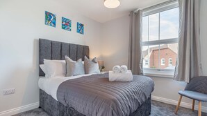 A neatly made bed with a grey headboard and pillows, a small bedside table with a lamp, and a window with curtains. - Host Stay The Bull Inn Flat 1 (Worcester)