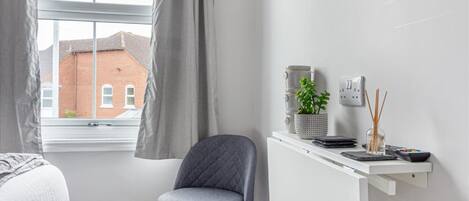 A room with a bed, a desk with a plant, and a view of a brick building outside.
