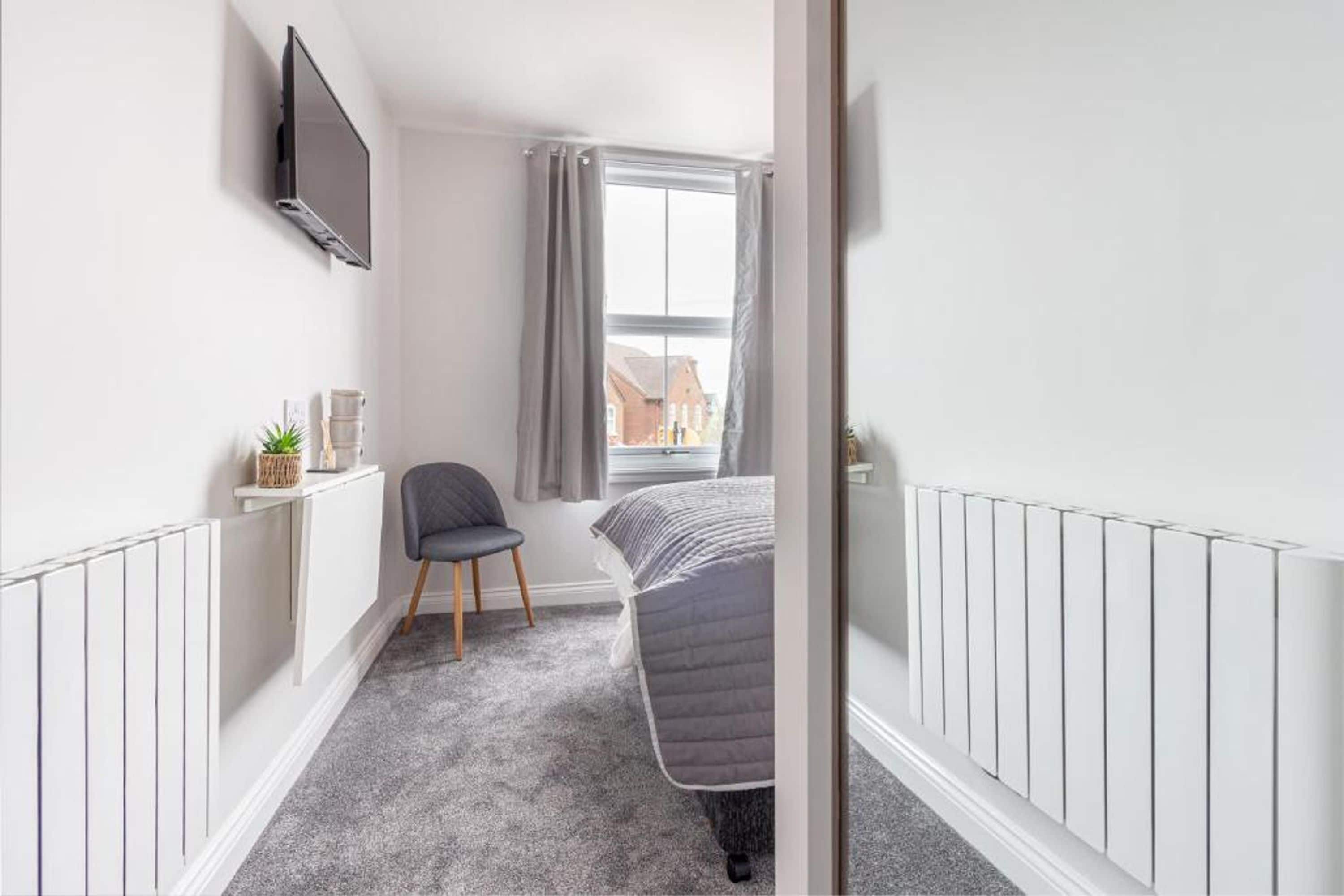A narrow room with a bed, a chair, a TV mounted on the wall, and a window with curtains.