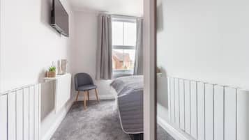 A narrow room with a bed, a chair, a TV mounted on the wall, and a window with curtains.