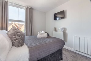 Una cama bien hecha con colcha gris, un almohadón y dos toallas enrolladas. Un televisor de pantalla plana está montado en la pared sobre una pequeña mesa blanca con un hervidor y una taza.