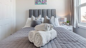 Un lit bien fait, avec une couverture matelassée grise et des serviettes blanches pliées, une tête de lit rembourrée grise, une petite table de chevet avec une lampe et une plante, et une œuvre d’art florale aux couleurs vives accrochée au mur.