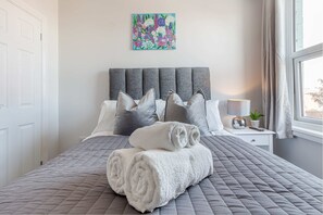 Ein ordentlich bezogenes Bett mit einer grauen gesteppten Tagesdecke und aufgerollten weißen Handtüchern, ein grau gepolsterter Kopfteil, ein kleiner Nachttisch mit Lampe und Pflanze sowie ein farbenfrohes Blumenbild an der Wand.