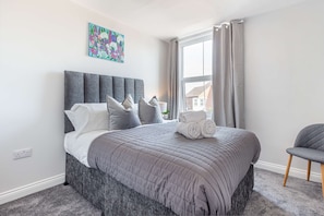 Una cama bien hecha con colcha gris, cabecera tapizada en gris y una obra de arte floral colorida en la pared.