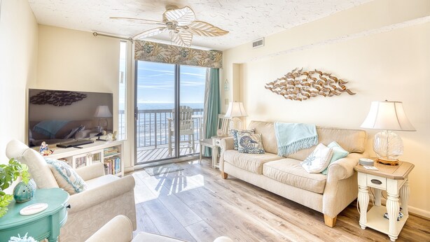 A spacious living room with a large window, a ceiling fan, and a balcony with a view. - Waterpointe II 705 | Oceanfront 3 Bedroom Condo (North Myrtle Beach)