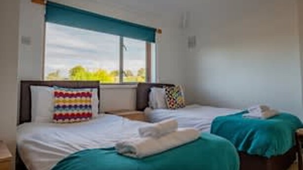 Ein Zimmer mit zwei Betten, jedes mit einer andersfarbigen Decke, einem Fenster mit Blick auf Grünflächen und einem farbenfrohen Kissen auf einem Bett.