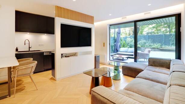 A modern kitchen with a flat-screen TV, a dining area with a table and chairs, and a large sofa in a bright living room with a sliding glass door to a patio. - Chic Apt With Garden - 1bd/2p - Neuilly-sur-seine (Neuilly-sur-Seine)