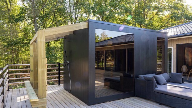 Een modern terras buiten met een houten pergola, een glazen omheining en een zithoek met kussens.