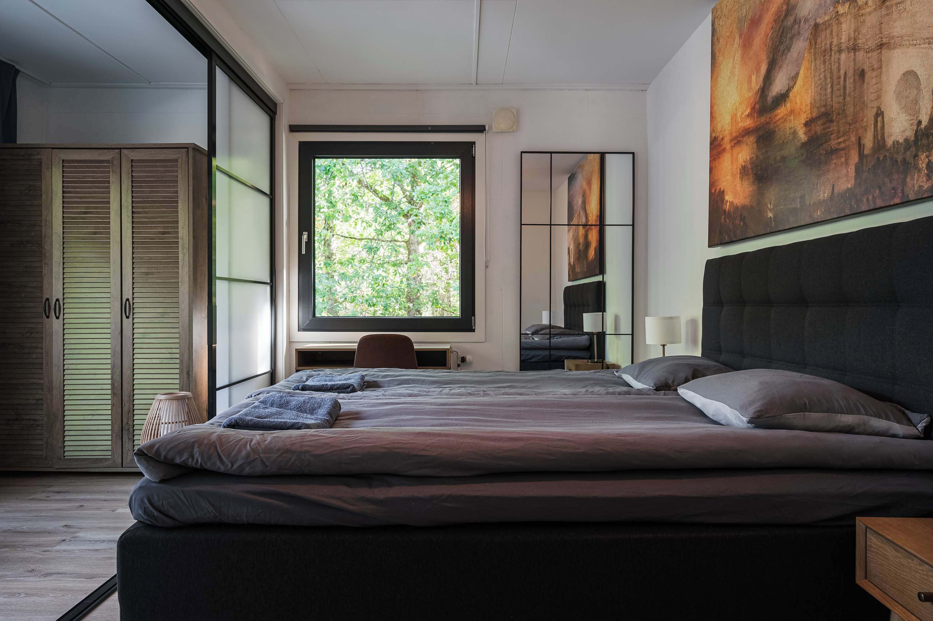 A modern bedroom with a large bed, a desk, and a window with a view of greenery.