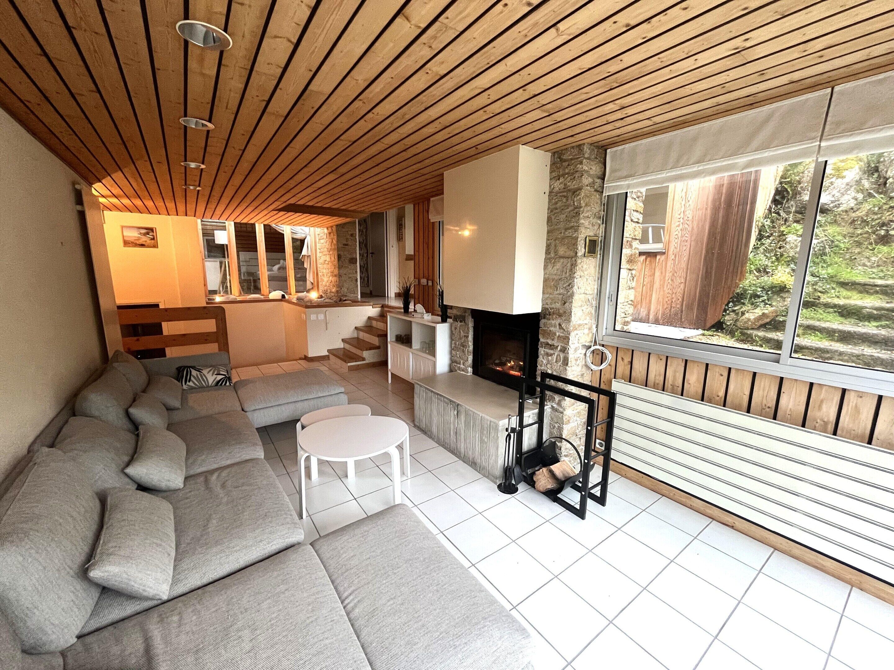 Un salon moderne doté d’une cheminée en pierre, d’un plafond en bois et d’une grande fenêtre donnant sur un jardin.
