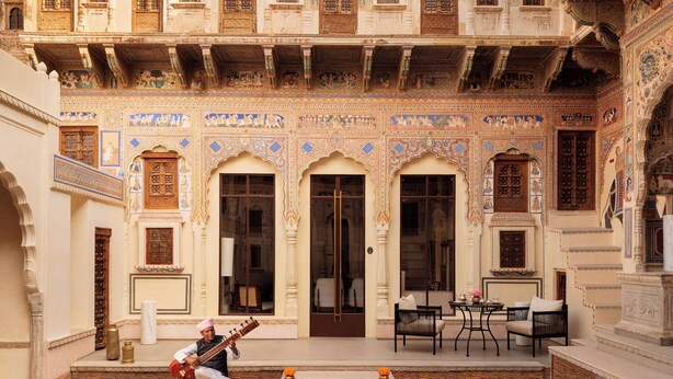 Un patio con detalles arquitectónicos intrincados, un músico tocando un instrumento de cuerda, y un área de descanso con sillas y una mesa.