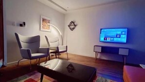 Ein modernes Wohnzimmer mit einem wandmontierten Flachbildfernseher, zwei Sesseln, einem Couchtisch und einem gemusterten Teppich.