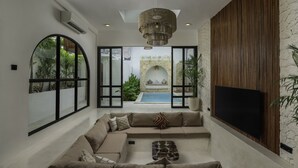 A modern living room with a large sectional sofa, a flat-screen TV mounted on a wooden wall, and a view of a swimming pool through large windows.