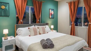 Un lit bien fait, avec des oreillers et une couverture, deux tables de chevet avec des lampes, une fenêtre avec des rideaux et un mur orné d’un tableau encadré.