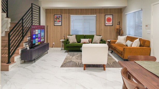 Un salon moderne avec un canapé vert, un canapé de couleur moutarde, un pouf blanc et une table à manger en bois. On y trouve un téléviseur posé sur un meuble, un escalier avec une rampe noire et un lambris en bois sur les murs.