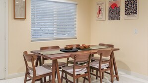 Un coin repas avec une table en bois, six chaises et une fenêtre avec des stores. Trois œuvres d’art encadrées ornent le mur.