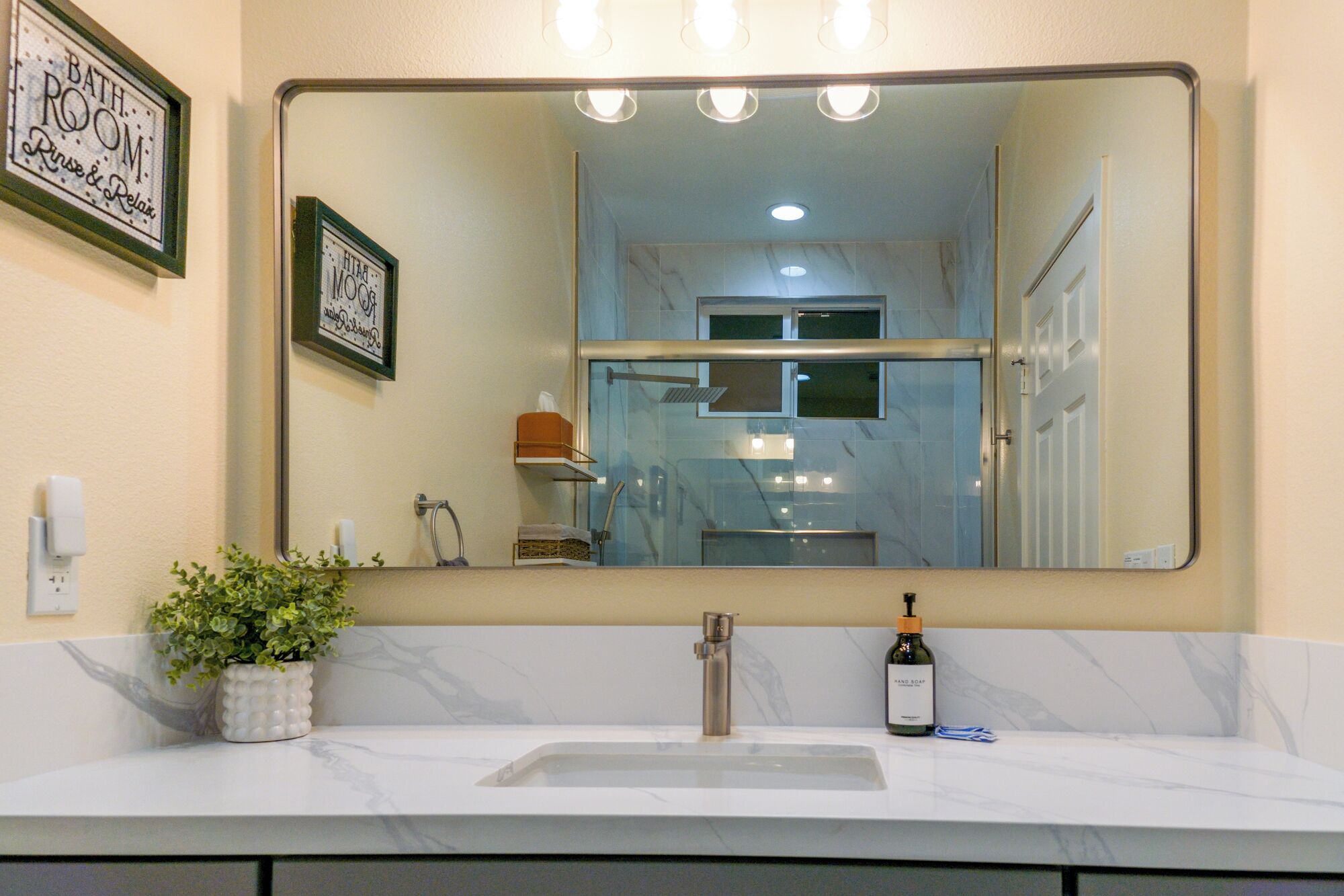 A bathroom with a large mirror, a sink, a potted plant, and a bottle of hand soap.