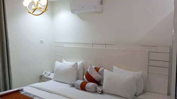 A modern hotel room with a large bed, white bedding, a bedside table, and a wall-mounted air conditioner. - Appleville Suites (Lagos)