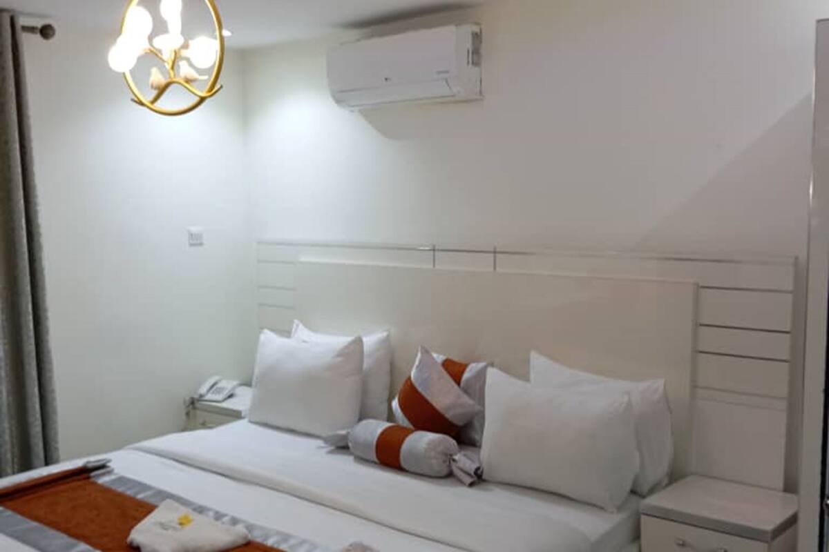 A modern hotel room with a large bed, white bedding, a bedside table, and a wall-mounted air conditioner.