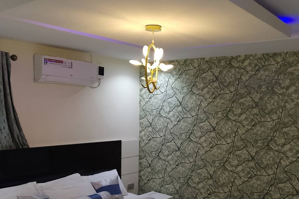 A modern bedroom with a large bed, a chandelier, a wall-mounted air conditioner, and a textured wall.