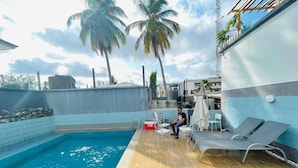 A pool area with a wooden deck, lounge chairs, and a person sitting by the pool. - May Shorlet Suites  (Abuja)