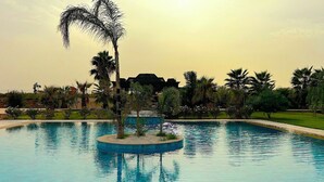 A large swimming pool with clear blue water, surrounded by palm trees and a building in the background. - Palais Bahida (marrakech)