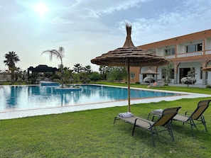 A swimming pool with lounge chairs and a thatched umbrella, surrounded by greenery and a building with a balcony.