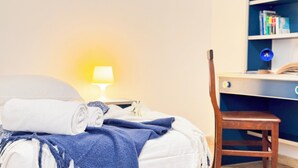Un letto rifatto con cura, un asciugamano bianco arrotolato, una coperta blu e un fiore bianco. Accanto al letto, una sedia di legno, un comodino con una lampada e una libreria con libri e un oggetto blu.