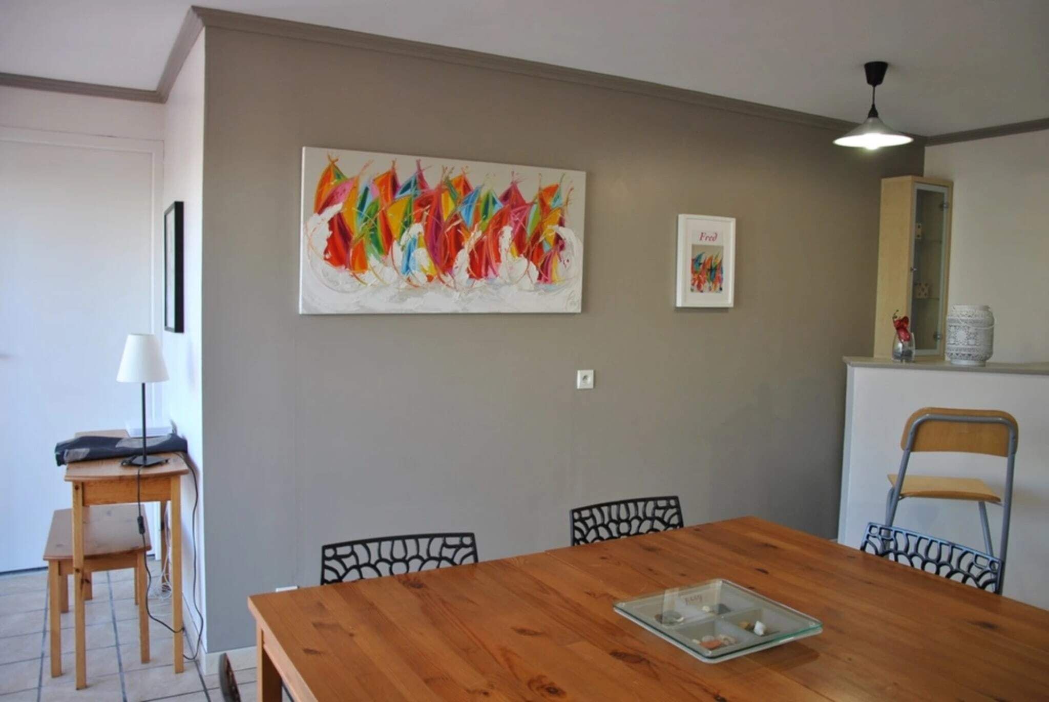 Un coin repas avec une table en bois, des chaises noires, un ventilateur de plafond et un tableau abstrait aux couleurs vives accroché au mur.