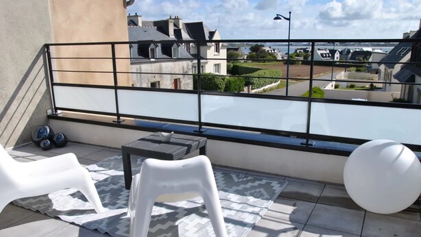 Een balkon met een metalen hekwerk, twee witte plastic stoelen, een klein rond tafeltje en een witte bolvormige object.