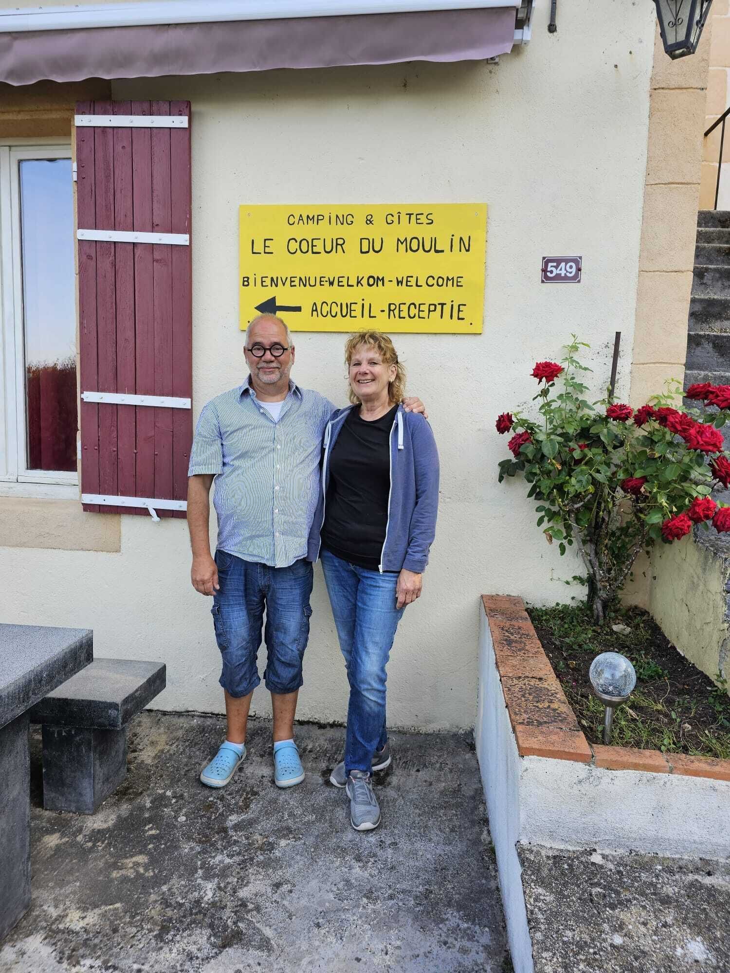 A couple standing in front of a building with a sign indicating camping and gîtes, welcoming visitors.
