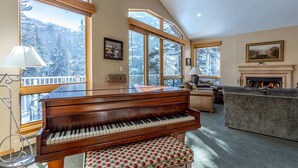 A spacious living room with a grand piano, large windows offering a view of a snowy landscape, a fireplace, and a cozy seating area. - Gore Creek Alpine Chalet (Vail)