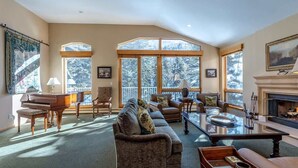 A spacious living room with a grand piano, comfortable seating, large windows, and a fireplace. - Gore Creek Alpine Chalet (Vail)