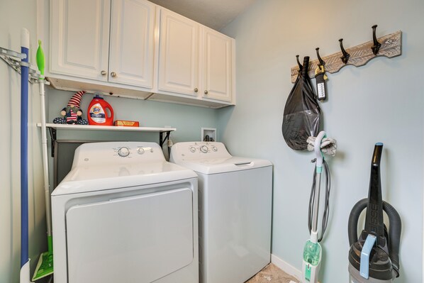 A laundry room with two front-loading washers and dryers, a shelf with cleaning supplies, and a vacuum cleaner on the wall. - Dock & Views: Lake Mitchell Home in Cadillac! (Cadillac)