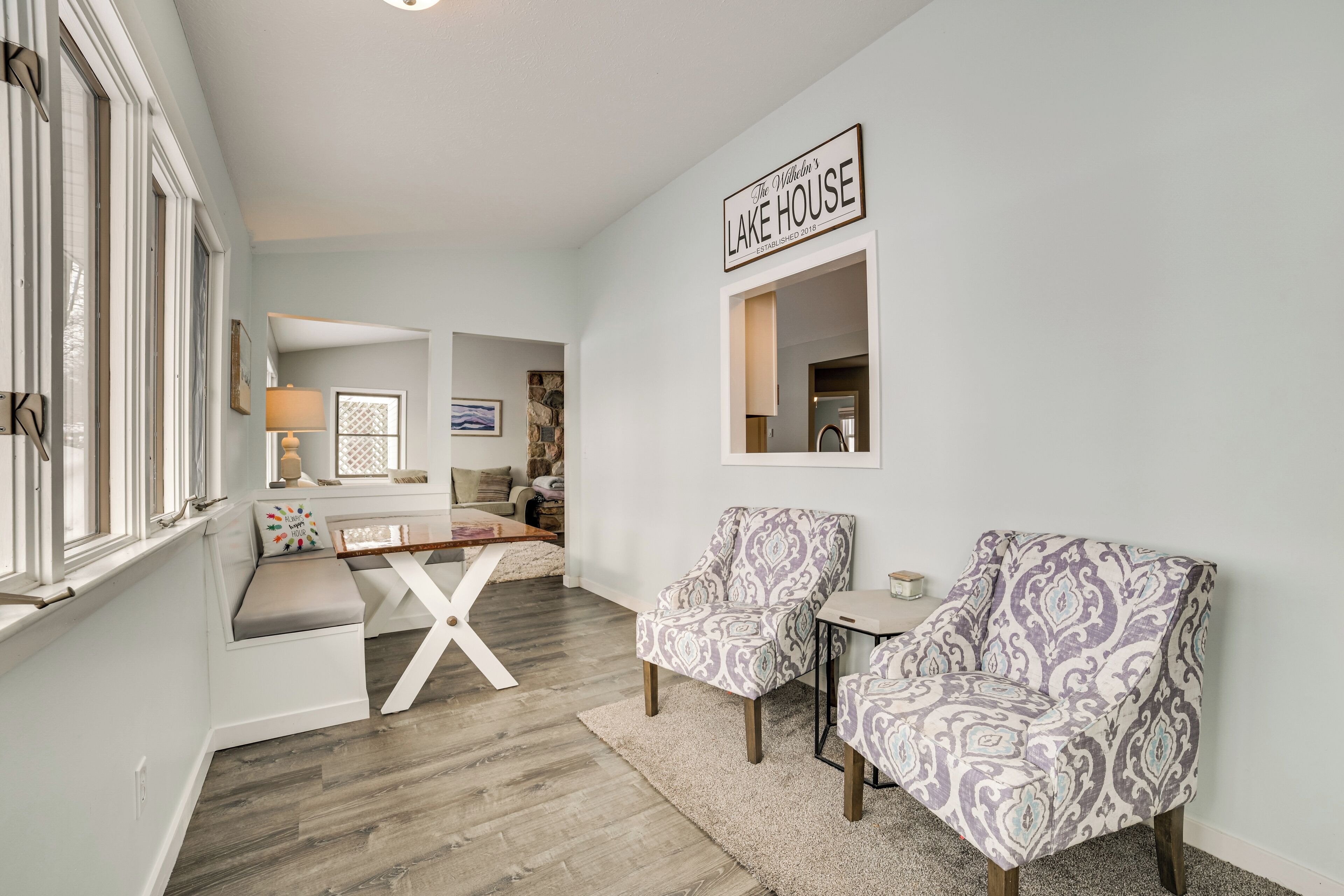 A modern living room with a white bench, two patterned armchairs, a small table, and a sign reading "The Wilkins' Lake House."