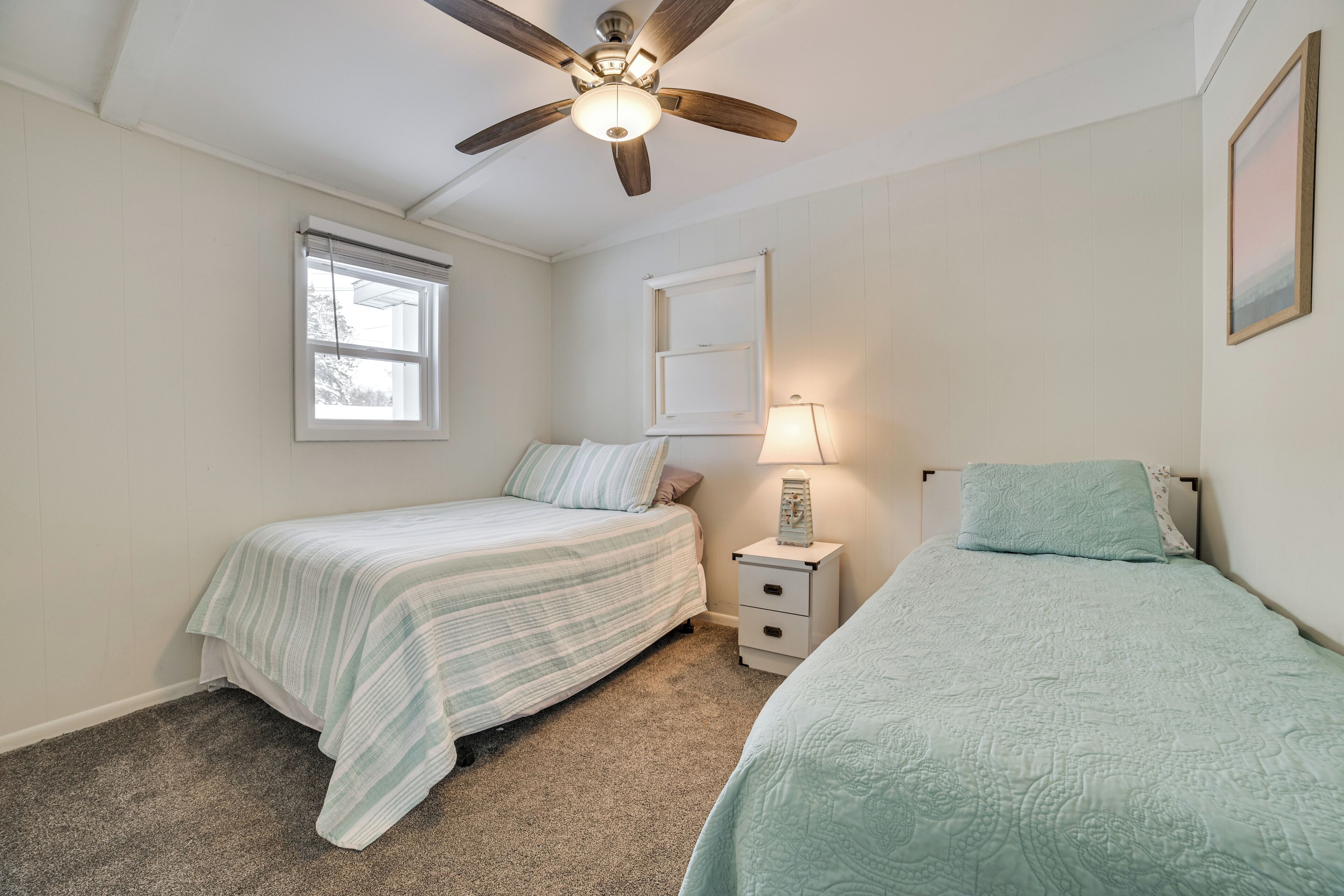 A bedroom with two beds, a ceiling fan, a window, a nightstand with a lamp, and a framed picture on the wall.