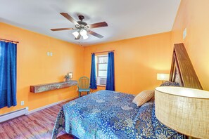 Un dormitorio con cama, ventilador de techo, ventana con cortinas, mesita de noche y lámpara.