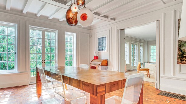 A spacious dining area with a large wooden table and clear plastic chairs, large windows with a view of greenery, and a modern pendant light fixture. - Hudson Valley Estate on 4 Acres w/ Private Pool! (Nyack)