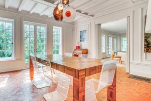 Un'ampia sala da pranzo con un grande tavolo in legno e sedie trasparenti in plastica, ampie finestre con vista su piante e un moderno lampadario a sospensione.