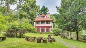 A two-story house with a red roof and white walls, surrounded by a lush garden with a stone pathway leading to a wooden bench area. - Hudson Valley Estate on 4 Acres w/ Private Pool! (Nyack)