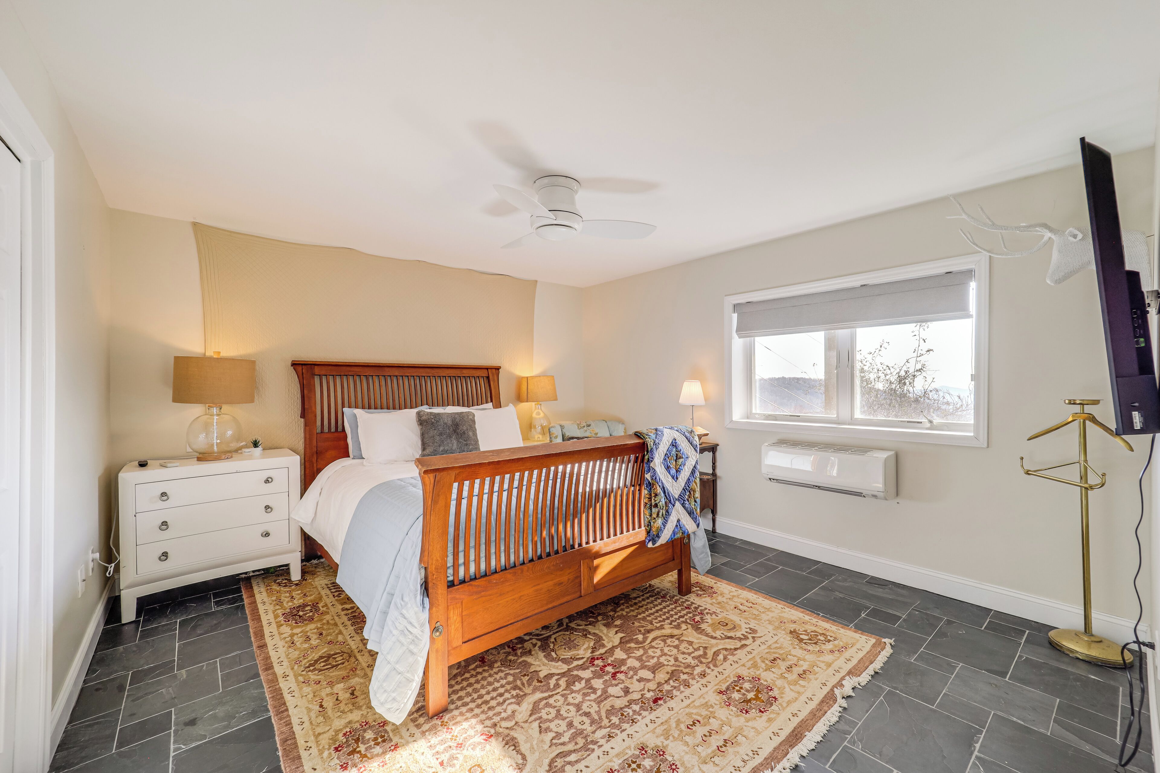A bedroom with a wooden bed, a ceiling fan, a nightstand with lamps, a window with a view, and a floor lamp.